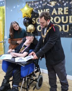 Mayor presents book to resident in wheelchair, and key worker stands at the side